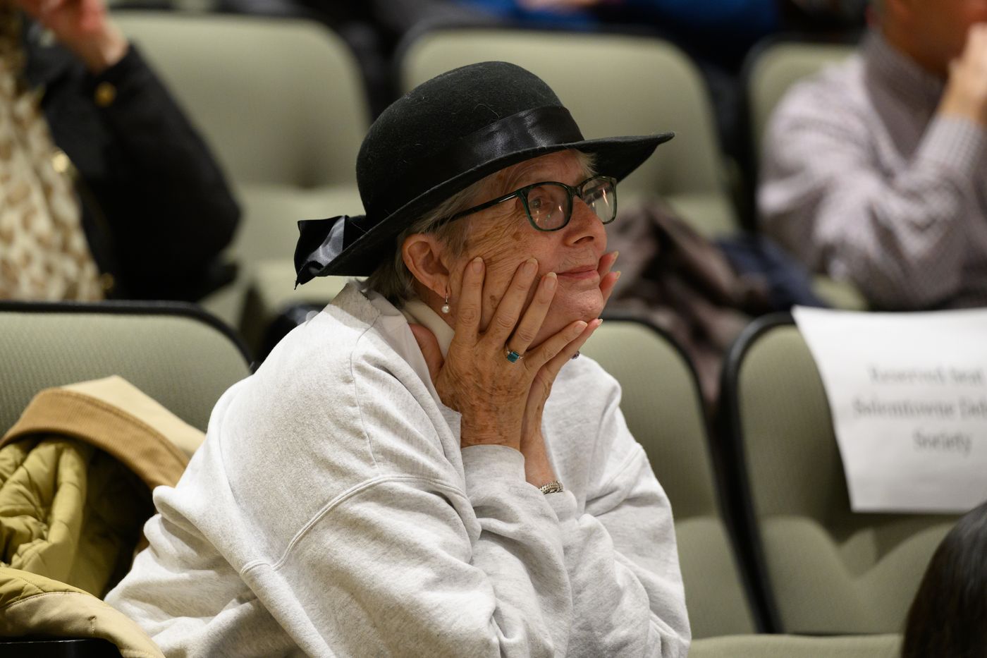 An audience member in a black hat rests her chin on her hands, listening intently.