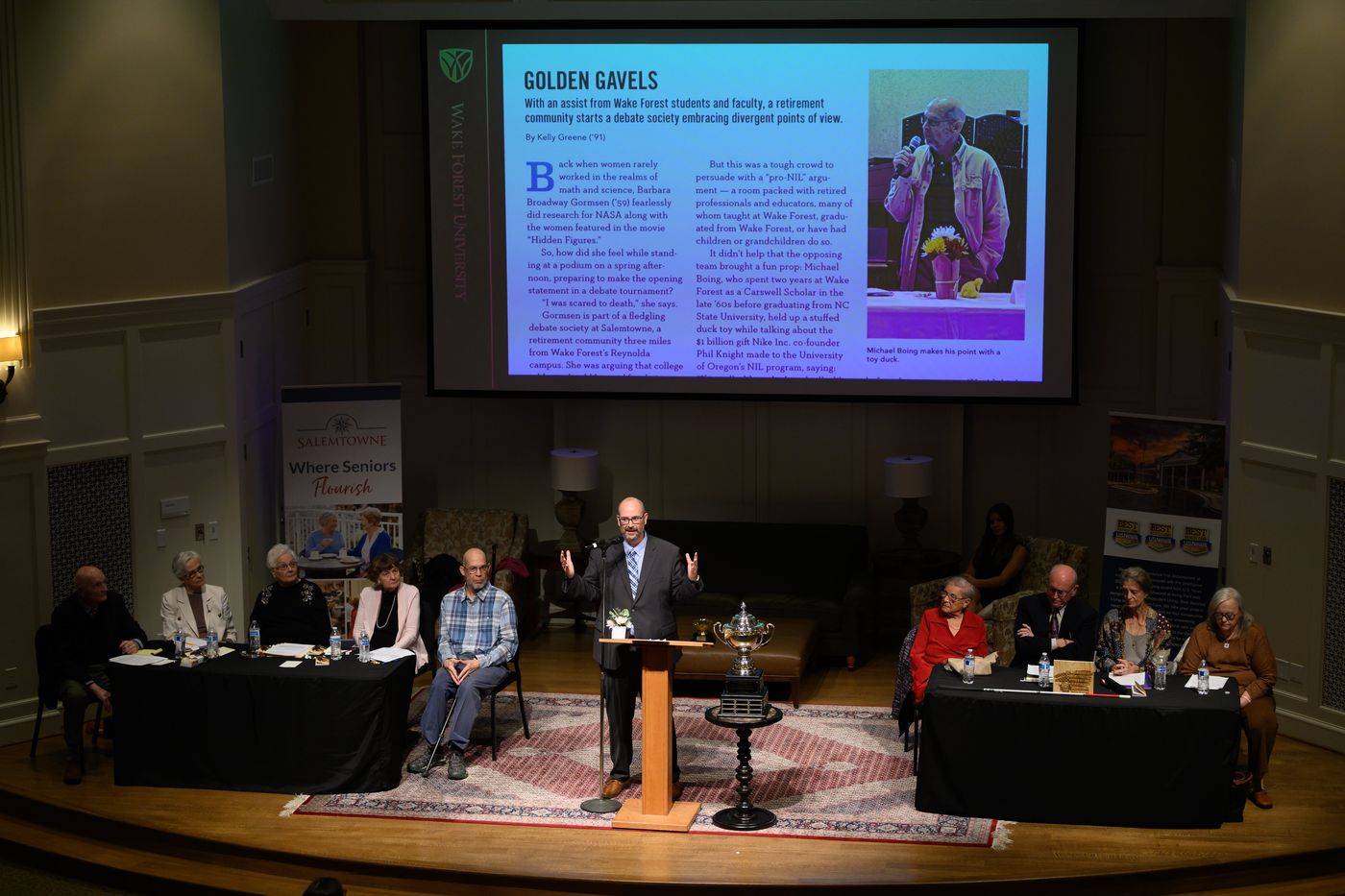 Wide view of the stage, with both teams seated at tables on either side of a podium and a projected magazine article behind.