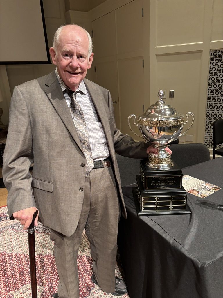 Dr. Allan Louden, smiling and holding a cane, stands beside the large silver championship trophy.